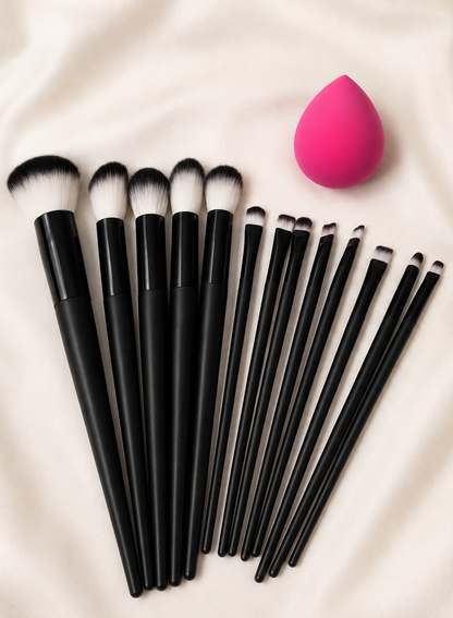 13-piece black makeup brush set with pink blending sponge, lined up on cream fabric